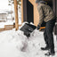 Uomo con giacca invernale e berretto grigio spalando neve con una pala nera fuori da una casa moderna in una giornata invernale. Scarpe da neve nere e guanti. Ambiente innevato con alberi e recinzioni in legno sullo sfondo.