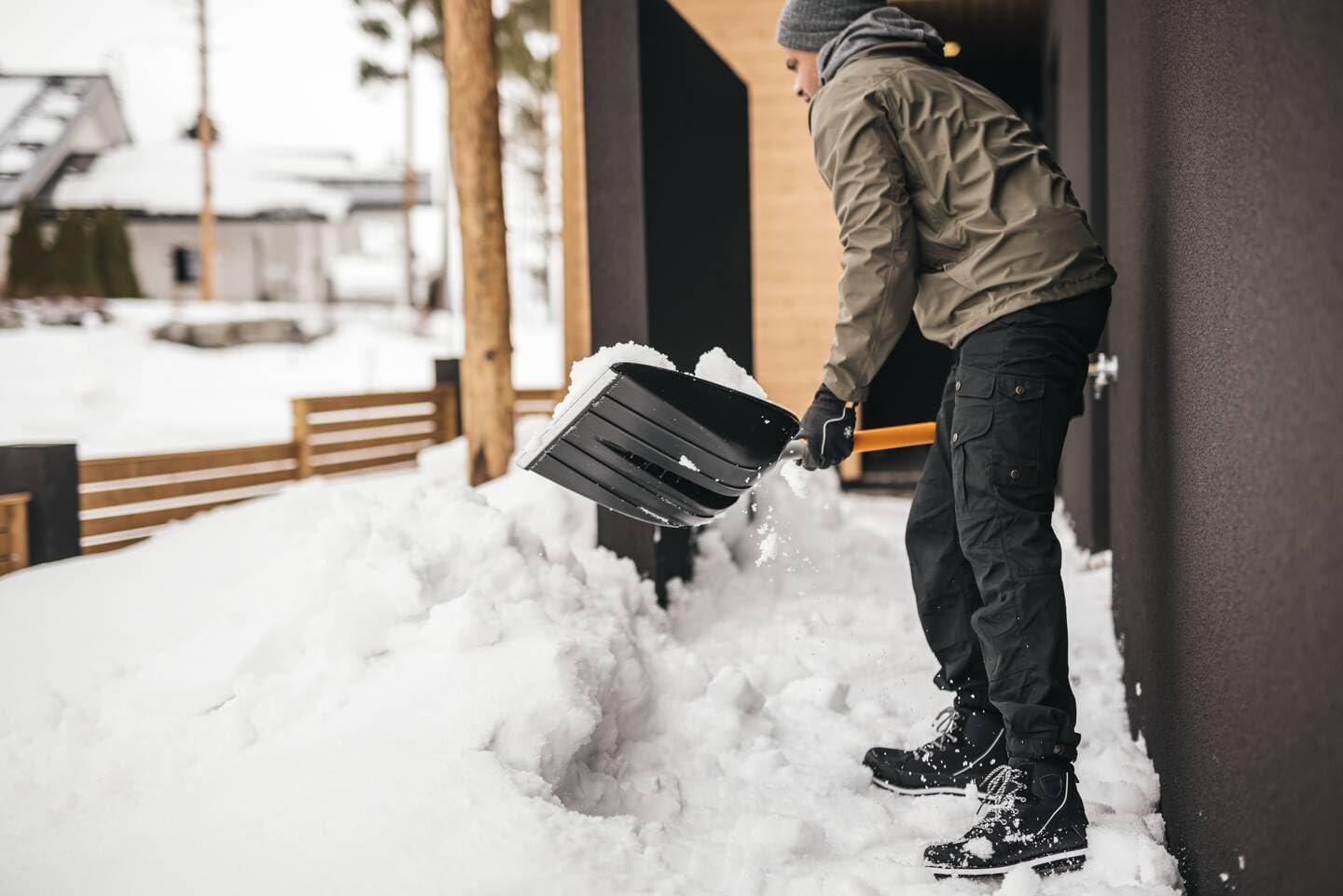 Uomo con giacca invernale e berretto grigio spalando neve con una pala nera fuori da una casa moderna in una giornata invernale. Scarpe da neve nere e guanti. Ambiente innevato con alberi e recinzioni in legno sullo sfondo.