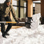 Uomo che spalando neve con pala ergonomica nera e manico arancione, indossando giacca verde, pantaloni neri, guanti e berretto grigio, in un ambiente domestico moderno con grandi finestre e mobili da esterno, durante una giornata invernale.