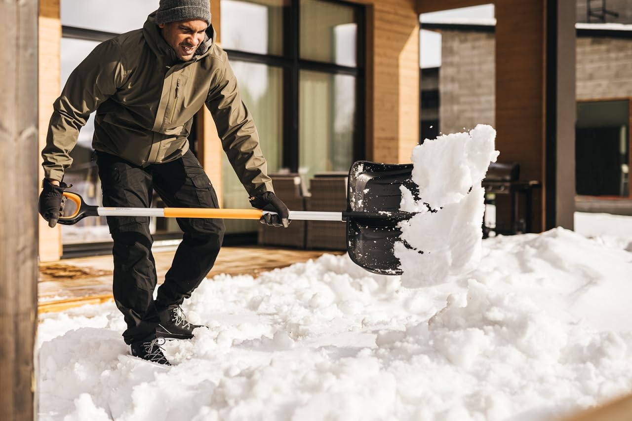 Uomo che spalando neve con pala ergonomica nera e manico arancione, indossando giacca verde, pantaloni neri, guanti e berretto grigio, in un ambiente domestico moderno con grandi finestre e mobili da esterno, durante una giornata invernale.