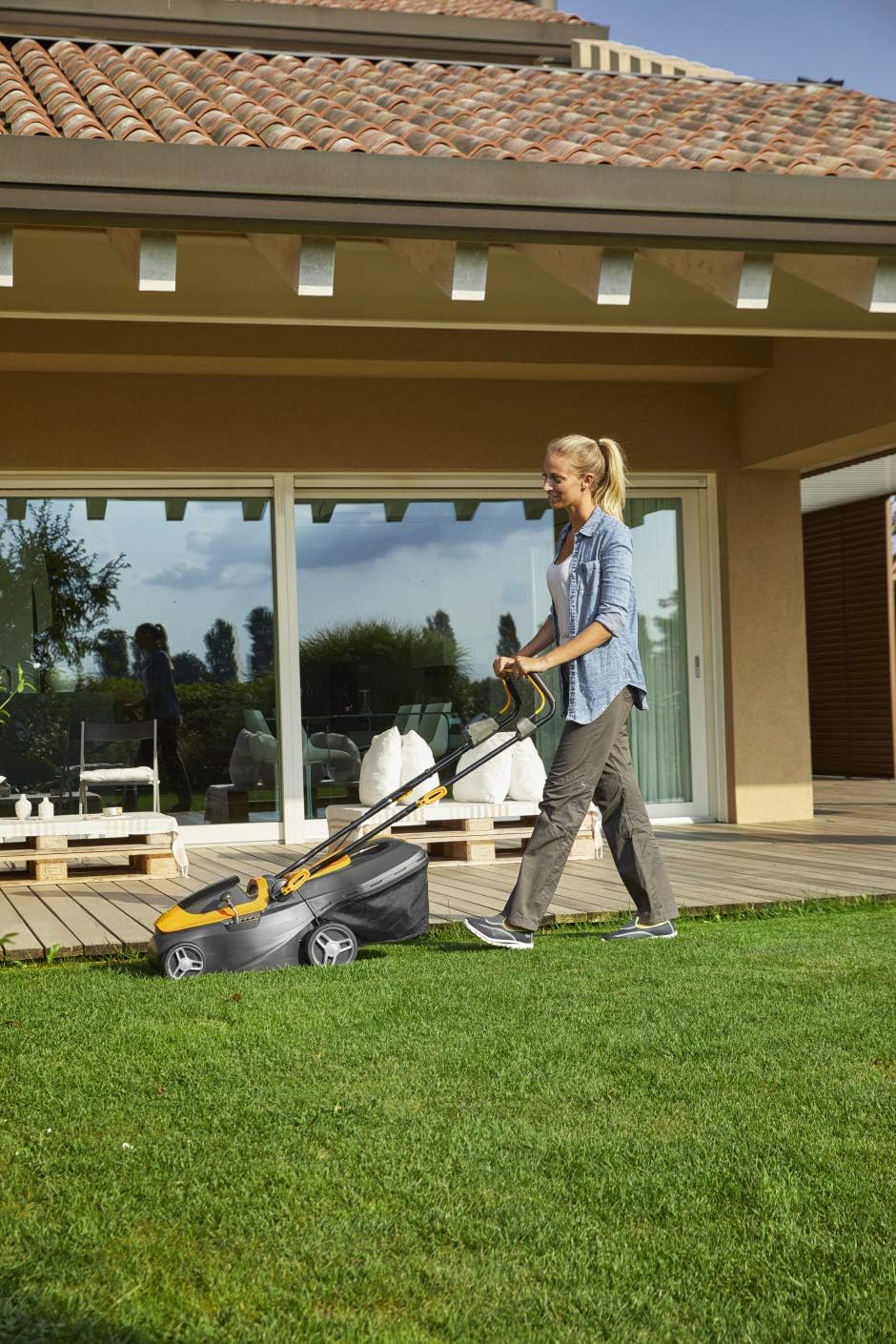 Donna che utilizza un tosaerba elettrico su un prato verde di fronte a una casa moderna con ampie finestre. Ideale per il giardinaggio domestico, il tosaerba offre praticità e efficienza nella cura del prato.