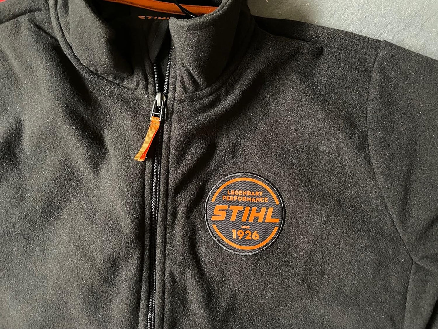 Giacca in pile nera STIHL con zip, dotata di logo arancione "Legendary Performance STIHL Since 1926" sul petto. Ideale per lavoro all'aperto e tempo libero, offre comfort e calore. Abbigliamento ufficiale STIHL di alta qualità.