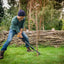 Uomo in giardino che utilizza una zappa per lavorare il terreno intorno a un albero, indossa cappello, guanti e stivali da lavoro. Sullo sfondo una staccionata di legno e alberi. Attrezzi da giardinaggio, cura del giardino, manutenzione esterna.