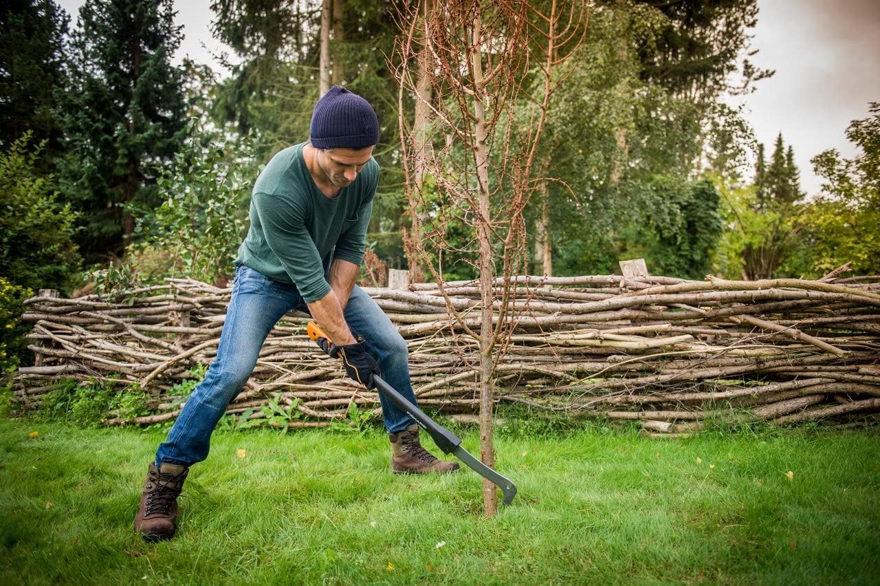 Uomo in giardino che utilizza una zappa per lavorare il terreno intorno a un albero, indossa cappello, guanti e stivali da lavoro. Sullo sfondo una staccionata di legno e alberi. Attrezzi da giardinaggio, cura del giardino, manutenzione esterna.