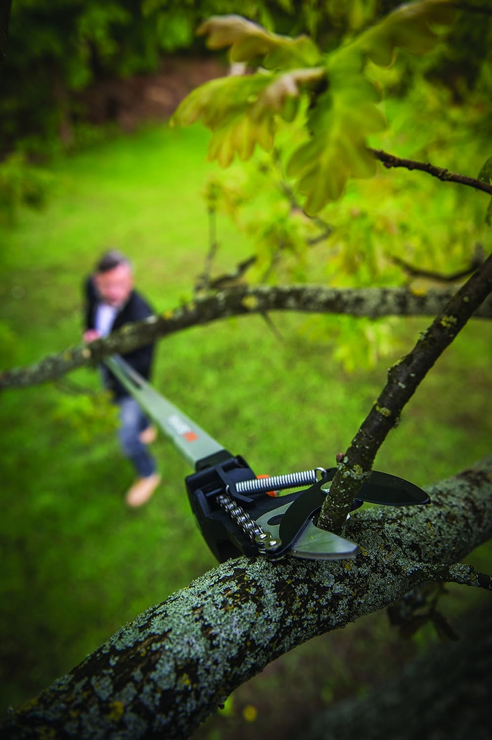 Attrezzo da giardinaggio per potatura telescopica in azione su un ramo di albero, utilizzato da una persona sfocata in sottofondo. Ideale per tagliare rami alti senza scala. Strumento professionale per cura del giardino, potatore telescopico preciso ed efficiente.