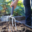 Persona che utilizza una forca da giardinaggio per lavorare il terreno in un giardino, con piante verdi sullo sfondo. Strumento da giardino ideale per aerare il suolo e prepararlo per la semina.