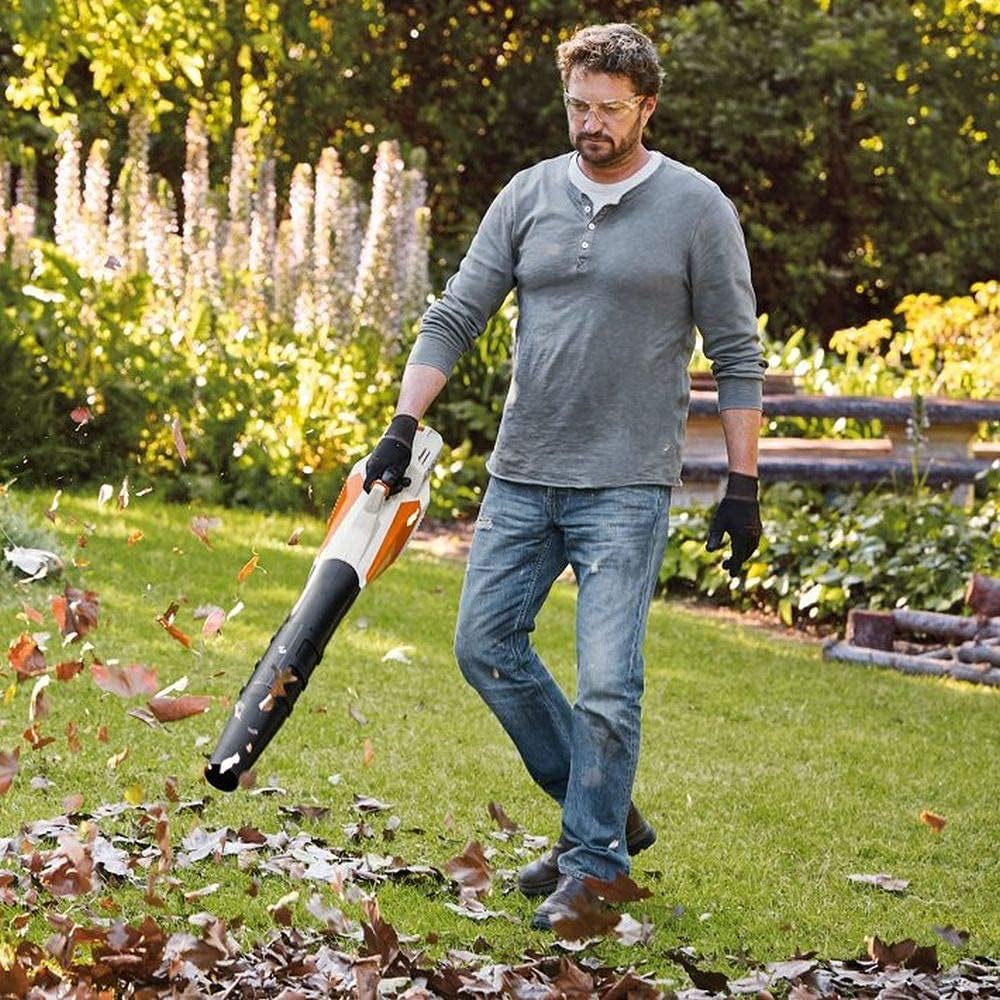 Uomo utilizza un soffiatore di foglie elettrico in un giardino verde, indossando occhiali protettivi, guanti neri, maglietta grigia e jeans. Attrezzo da giardinaggio potente per la pulizia del prato dalle foglie autunnali.