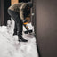 Uomo che spalando la neve con una pala ergonomica vicino a una casa moderna in inverno. Indossa abbigliamento invernale, inclusi giacca, pantaloni e stivali impermeabili. Scenario di montagna innevata. Strumenti per rimozione neve.