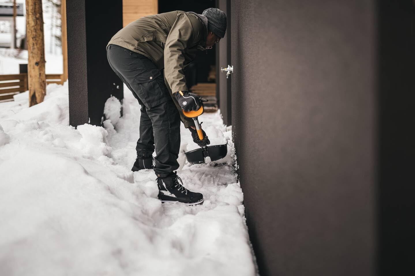 Uomo che spalando la neve con una pala ergonomica vicino a una casa moderna in inverno. Indossa abbigliamento invernale, inclusi giacca, pantaloni e stivali impermeabili. Scenario di montagna innevata. Strumenti per rimozione neve.