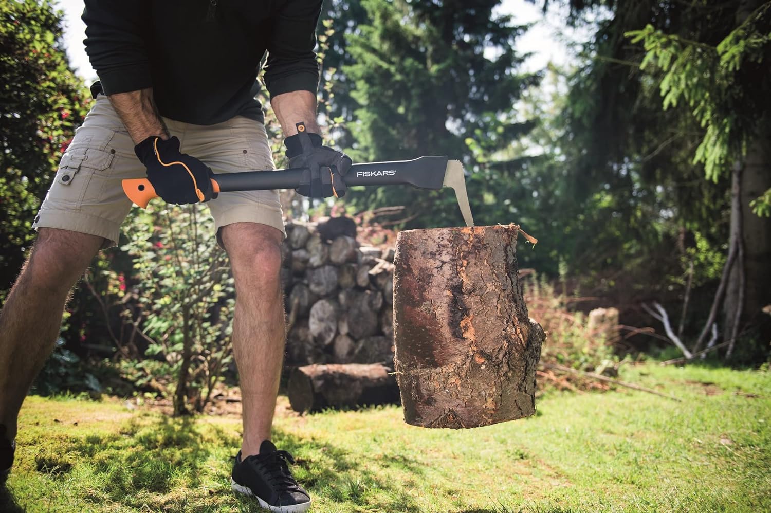 Persona che utilizza un'accetta Fiskars per spaccare un tronco di legno in un giardino. Indossa guanti di protezione e pantaloncini corti. Perfetta per lavori di giardinaggio e taglio della legna. Ambiente esterno con alberi e tronchi sullo sfondo.