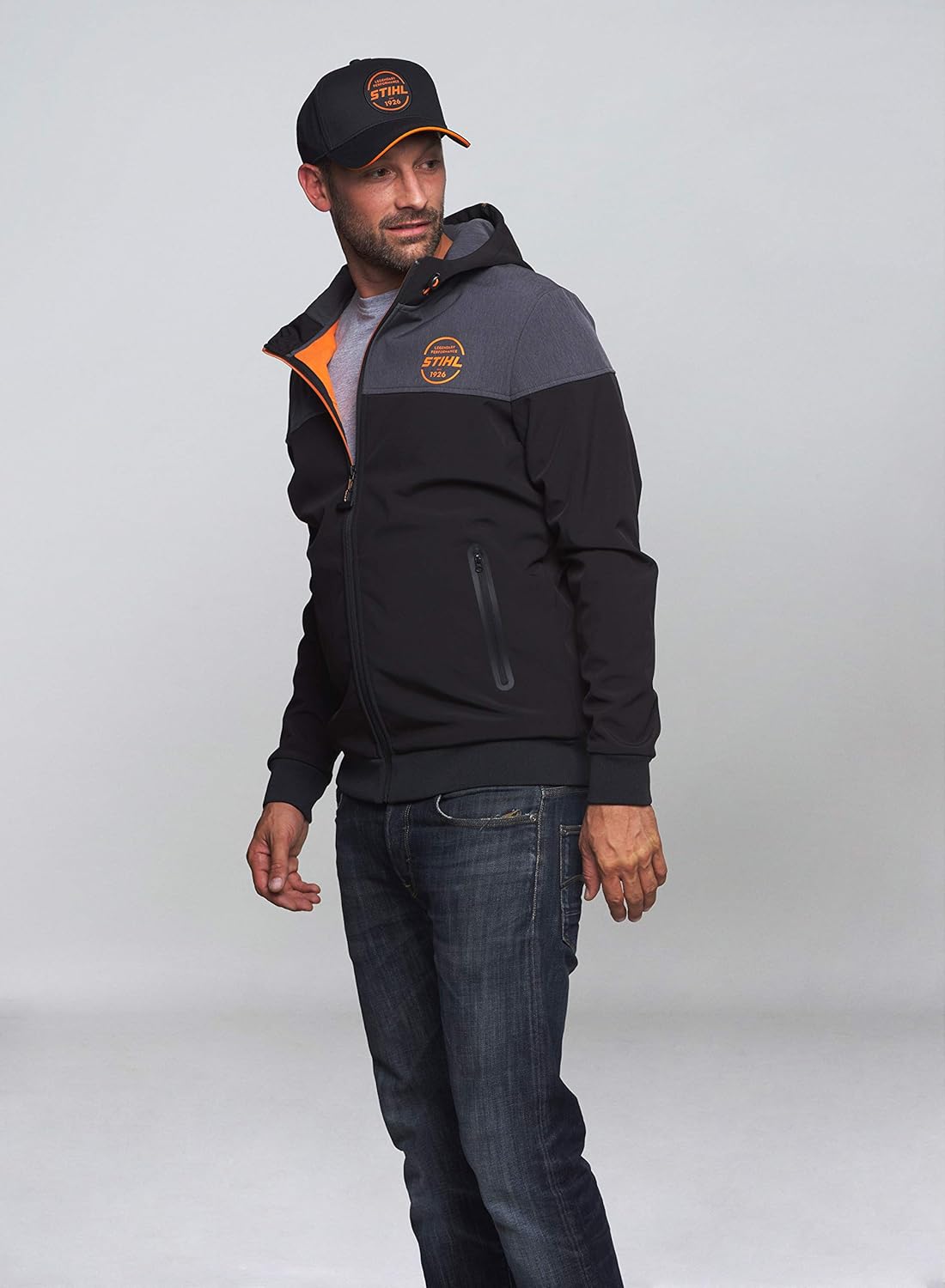 Giacca con cappuccio STIHL nera e grigia con zip, indossata da uomo con cappellino coordinato. Giacca sportiva, casual, perfetta per l'outdoor, con logo STIHL arancione sul petto. Abbigliamento uomo, moda autunnale.