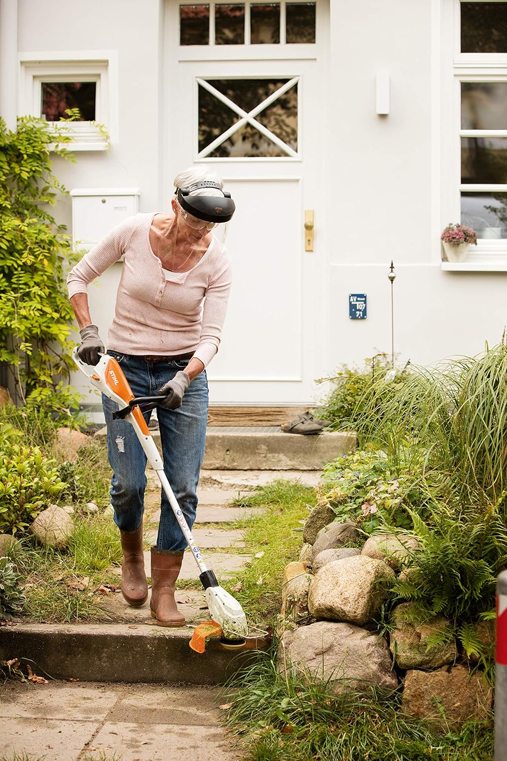 Donna che utilizza un decespugliatore Stihl per curare il giardino davanti alla casa. Indossa guanti, jeans e stivali per protezione. Ideale per lavori di giardinaggio e manutenzione del prato. Efficiente e pratico per uso domestico.