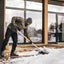 Uomo che spalare la neve su una terrazza di legno davanti a una casa moderna con grandi finestre. Indossa giacca invernale, guanti, pantaloni neri e stivali da neve. Attività invernale all'aperto, rimozione neve, manutenzione casa.