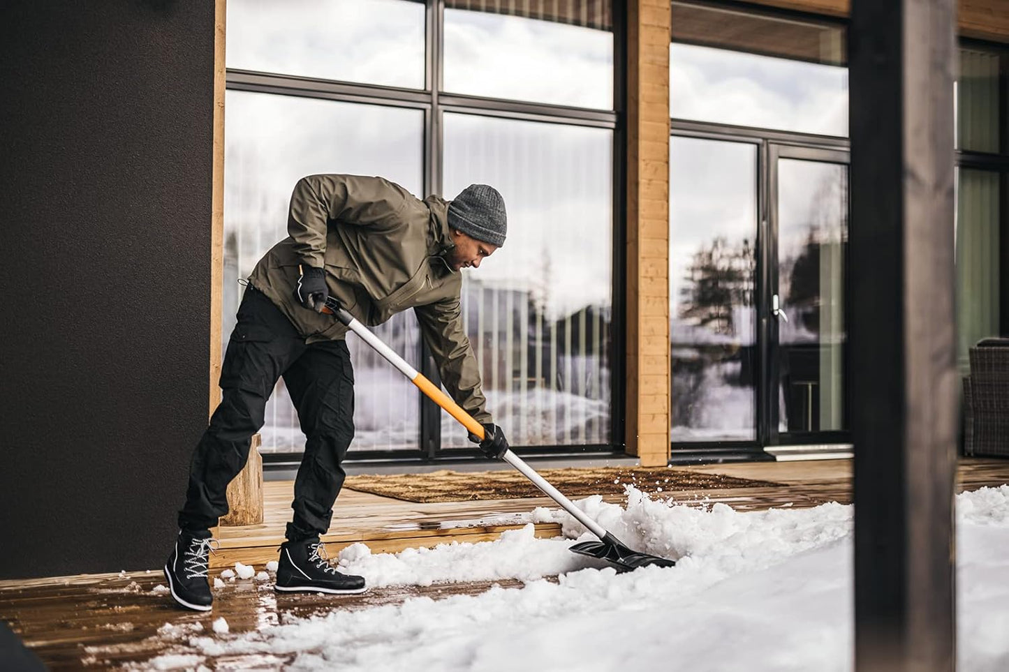 Uomo che spalare la neve su una terrazza di legno davanti a una casa moderna con grandi finestre. Indossa giacca invernale, guanti, pantaloni neri e stivali da neve. Attività invernale all'aperto, rimozione neve, manutenzione casa.