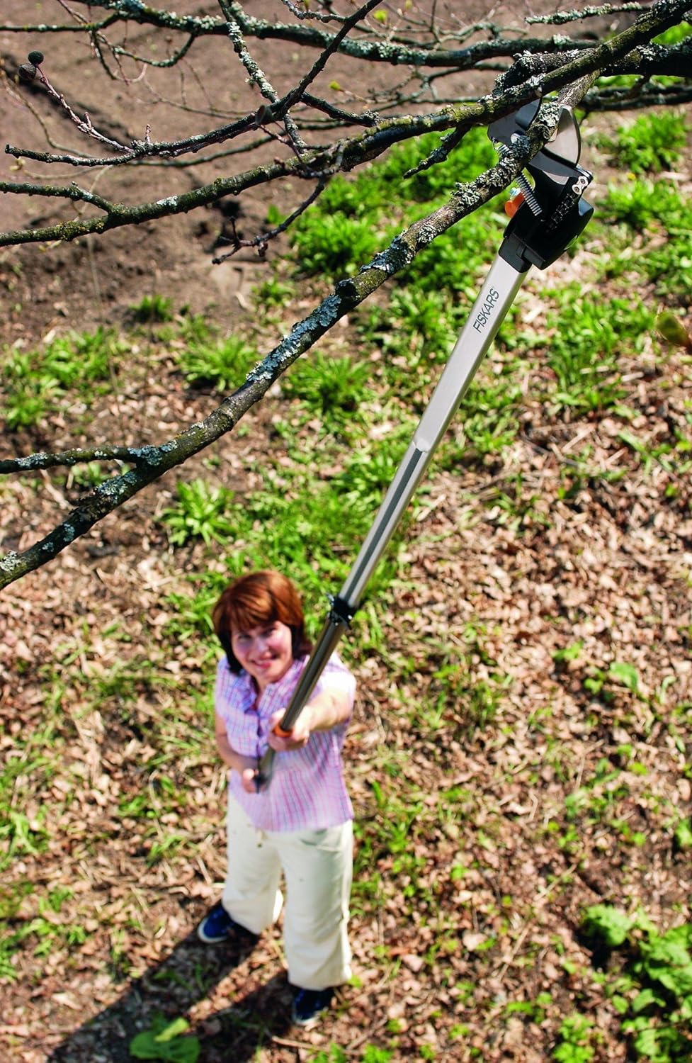 Donna che usa un potatore telescopico Fiskars per tagliare rami alti in giardino. Attrezzo da giardinaggio ergonomico e leggero per potatura precisa. Utensile ideale per la manutenzione di alberi e arbusti.