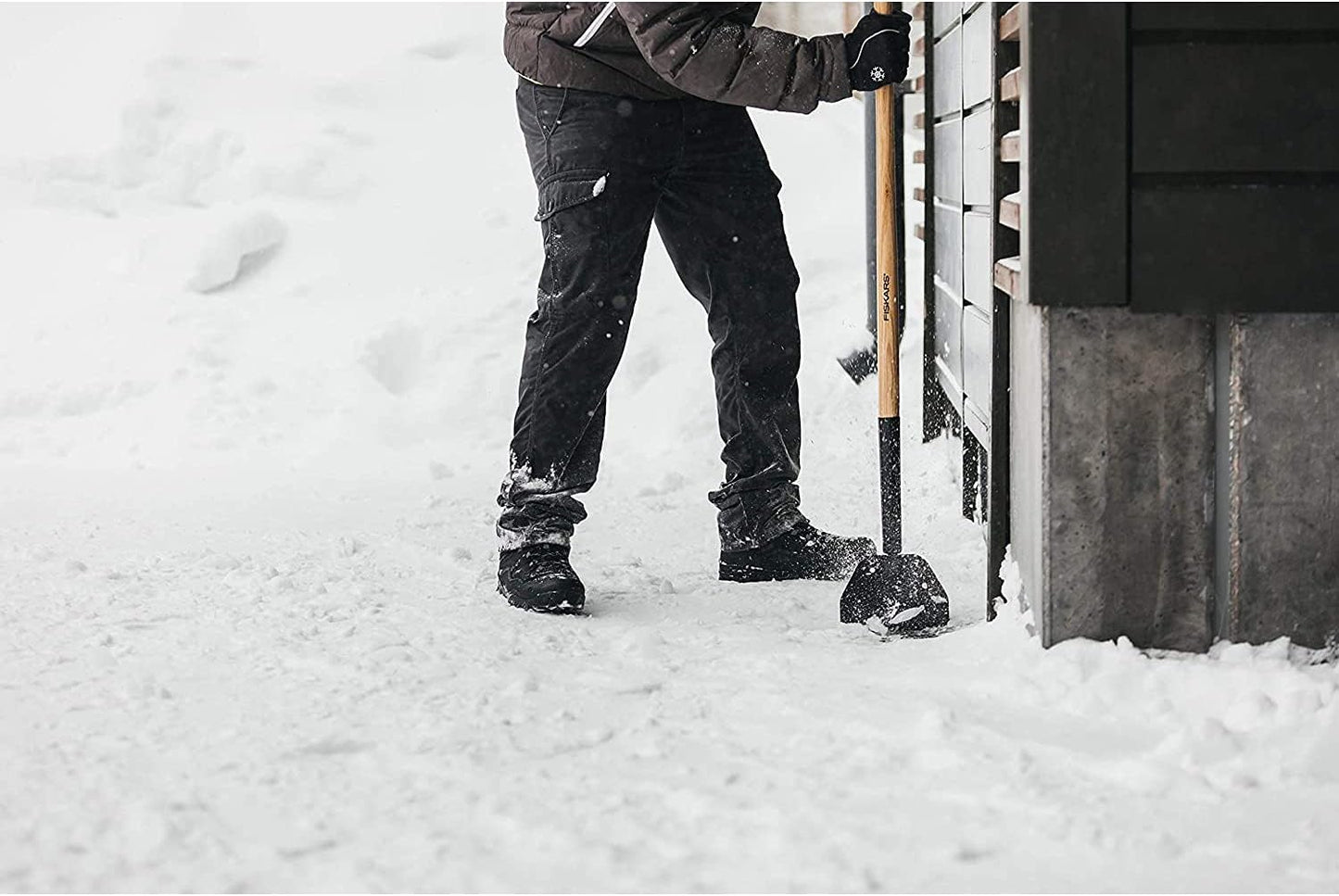 Persona che spalare neve con pala Fiskars, pantaloni neri e guanti invernali, davanti a una casa in legno. Strumento ideale per la rimozione della neve in inverno.