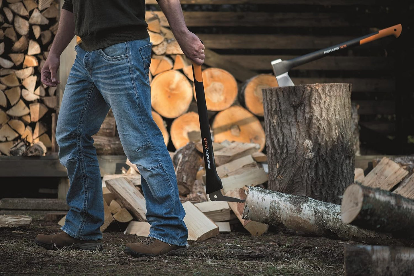 Uomo che indossa jeans e scarponi da lavoro, impugnando un'ascia Fiskars X27 mentre spacca legna in un cortile con tronchi tagliati sullo sfondo. Strumento ideale per il taglio di legna da ardere e lavori di giardinaggio.