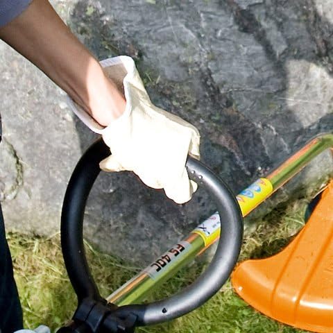 Persona con guanti da giardinaggio maneggia un decespugliatore arancione su prato verde, vicino a una roccia. Attrezzo da giardino per taglio erba e manutenzione aree verdi. Utensile da giardino per lavori di precisione.