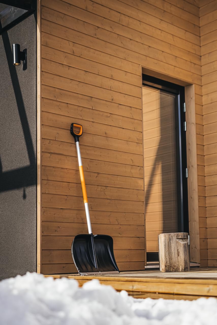 Pala da neve nera e arancione appoggiata a una parete di legno di una casa moderna. Strumento essenziale per spalare la neve durante l'inverno. Ambiente esterno con neve sul pavimento. Accessorio per la pulizia della neve.