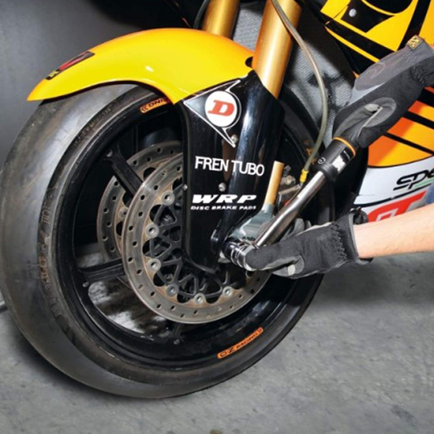 Immagine di una ruota anteriore di una moto con un copricerchio giallo. La ruota è montata con un disco freno con la scritta "Fren Tubo WRP Disc Brake Pads". Una mano con un guanto nero sta usando una chiave dinamometrica per lavorare sulla ruota.