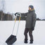 Uomo con giacca invernale e cappello di lana utilizza una pala da neve in plastica con manico in metallo per rimuovere la neve in un paesaggio innevato, circondato da alberi spogli e cielo grigio.