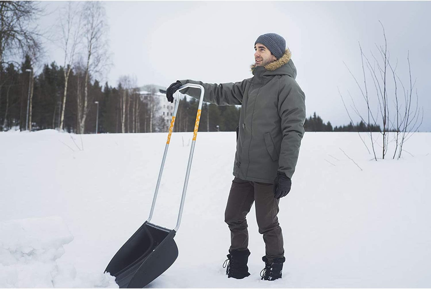 Uomo con giacca invernale e cappello di lana utilizza una pala da neve in plastica con manico in metallo per rimuovere la neve in un paesaggio innevato, circondato da alberi spogli e cielo grigio.