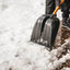 Persona che utilizza una pala da neve nera con manico giallo per rimuovere la neve da un ponte di legno. Indossa guanti, pantaloni invernali e stivali neri. Strumento ideale per spalare la neve durante l'inverno.