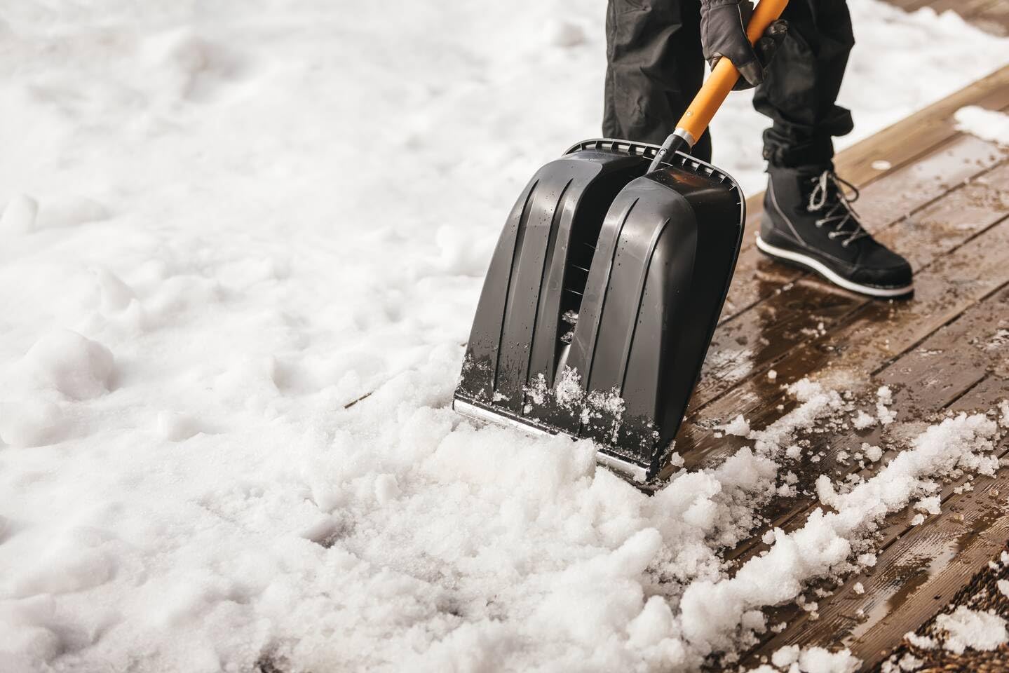 Persona che utilizza una pala da neve nera con manico giallo per rimuovere la neve da un ponte di legno. Indossa guanti, pantaloni invernali e stivali neri. Strumento ideale per spalare la neve durante l'inverno.