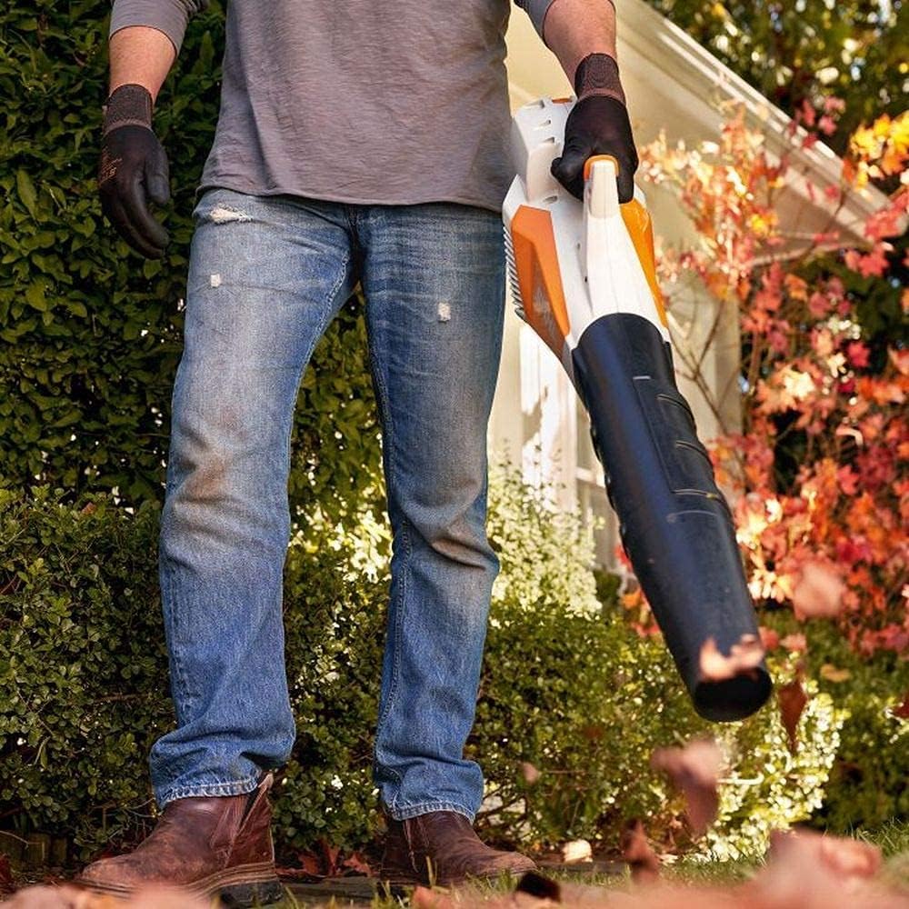 Uomo in jeans e stivali utilizza un soffiatore di foglie elettrico in giardino, rimuovendo foglie autunnali. Indossa guanti per protezione. Alberi e cespugli sullo sfondo. Strumento ideale per la pulizia esterna e la manutenzione del giardino.