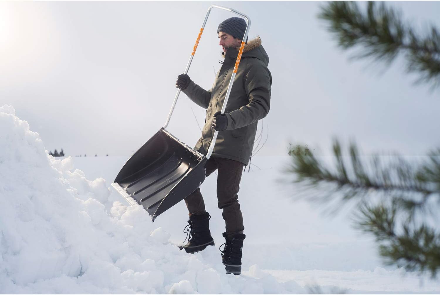 Uomo con giacca invernale e berretto usa una pala da neve ergonomica su un paesaggio innevato, attrezzatura ideale per rimuovere la neve in inverno, robusta e facile da maneggiare.