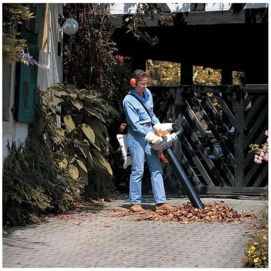 Persona che utilizza un soffiatore di foglie in un giardino, indossando cuffie protettive e guanti. Il soffiatore è ideale per la pulizia di foglie secche su vialetti e cortili. Ambiente esterno ben curato con piante e fiori.