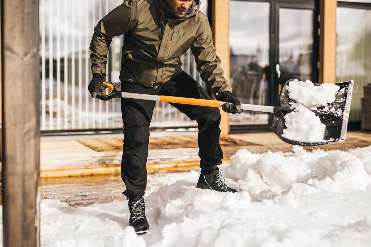 Uomo in giacca invernale e guanti neri che spalano la neve con una pala ergonomica su un patio di legno davanti a una casa moderna. Scarpe da neve robuste e pantaloni impermeabili. Preparazione per l'inverno e manutenzione esterna.