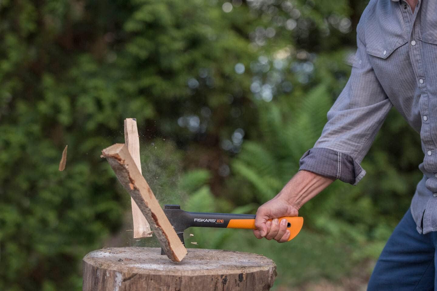 Persona che spacca legna con un'ascia Fiskars X7 su un ceppo di legno in un ambiente esterno, indossando una camicia grigia e jeans. Dettaglio dell'azione di spaccatura della legna con schegge che volano.