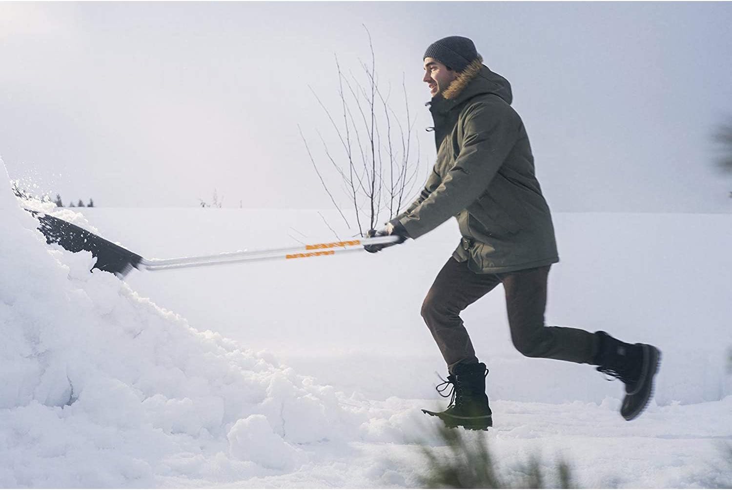 Uomo che spala neve con pala telescopica, vestito con giacca invernale, guanti, berretto e stivali, in un paesaggio innevato. Strumento ideale per rimuovere neve da vialetti e tetti durante l'inverno.