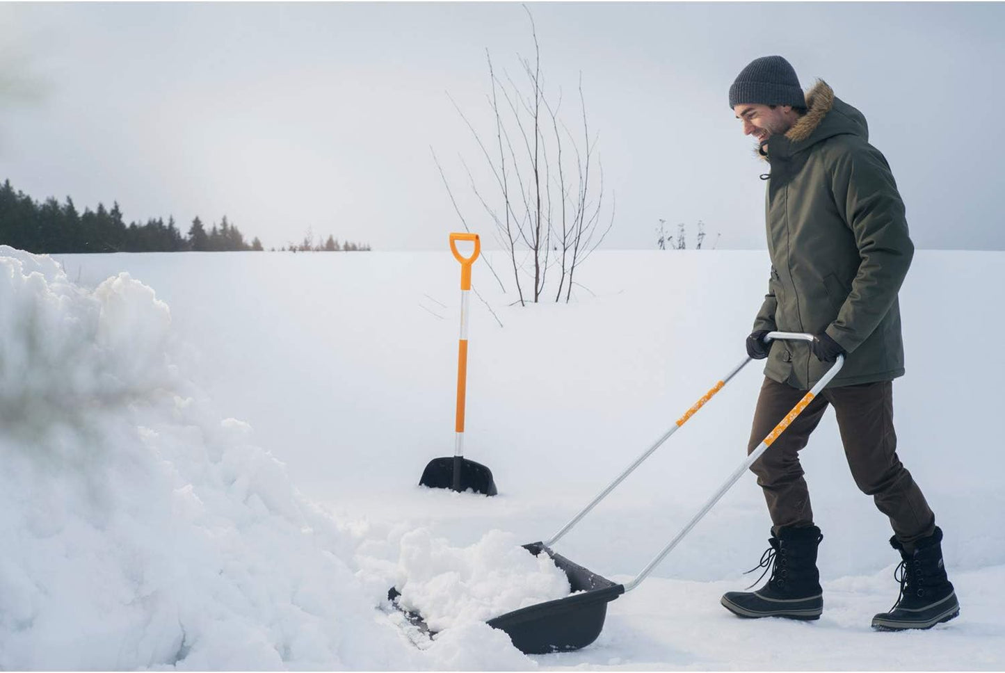 Uomo che spalare la neve con pala ergonomica invernale, vestito con giacca verde, pantaloni marroni e berretto grigio, in un paesaggio innevato con alberi sullo sfondo. Strumenti per spalare la neve.