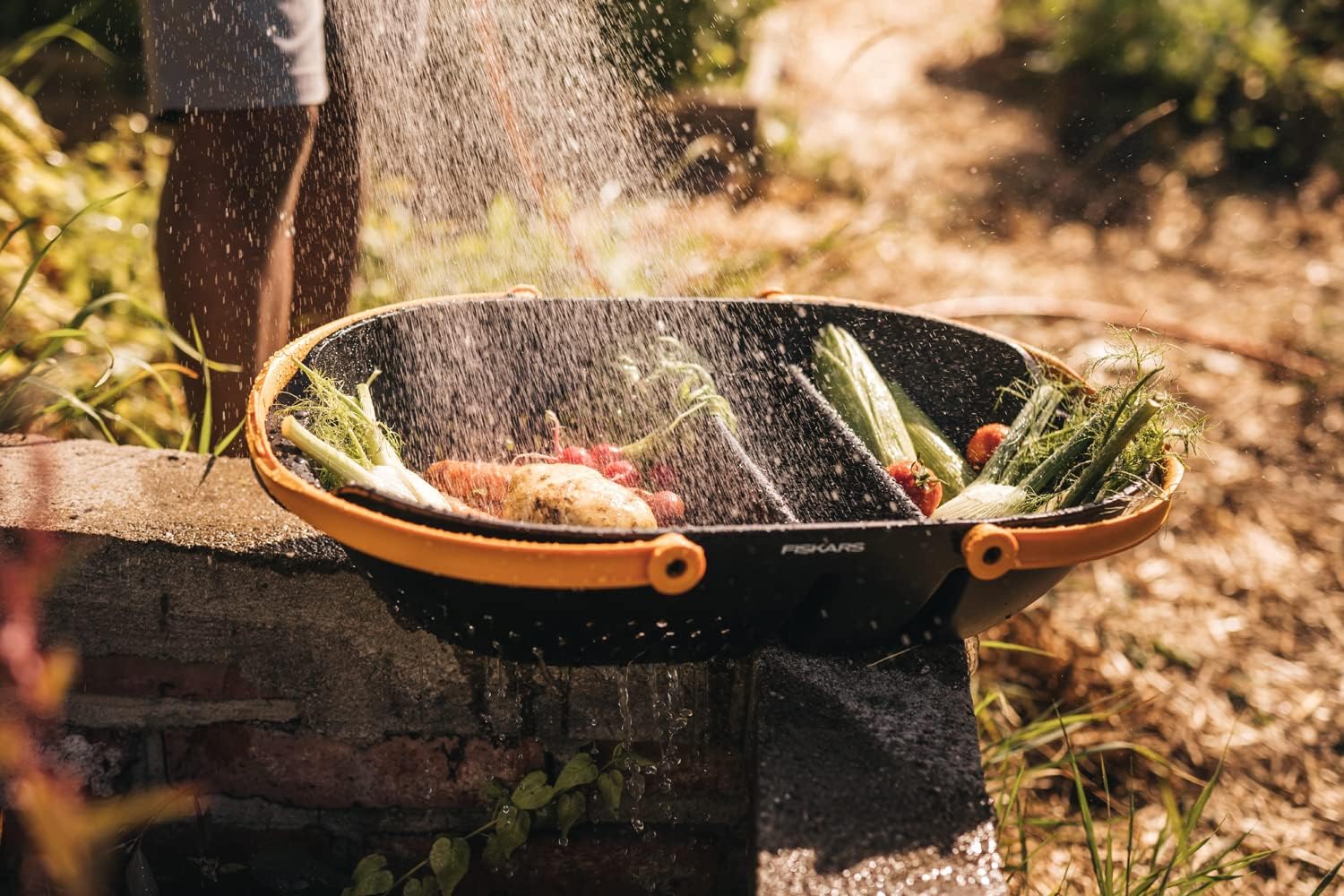 Colino da cucina Fiskars con manici arancioni, utilizzato per lavare verdure fresche come carote, cipolle e pomodori in un ambiente esterno soleggiato. Ideale per il giardinaggio e la preparazione di cibi sani.