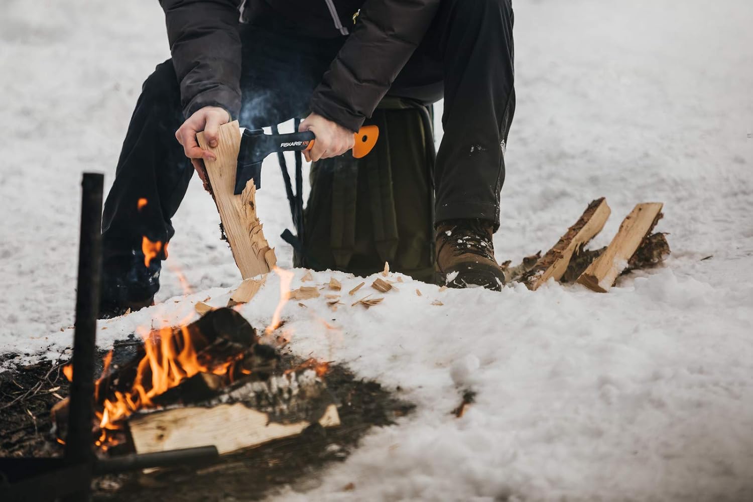 Persona che spacca legna con un'ascia Fiskars vicino a un fuoco da campo su neve, attrezzatura da campeggio invernale, abbigliamento outdoor, survivalismo, escursionismo invernale, preparazione del fuoco, attrezzi da campeggio, avventura nella natura.