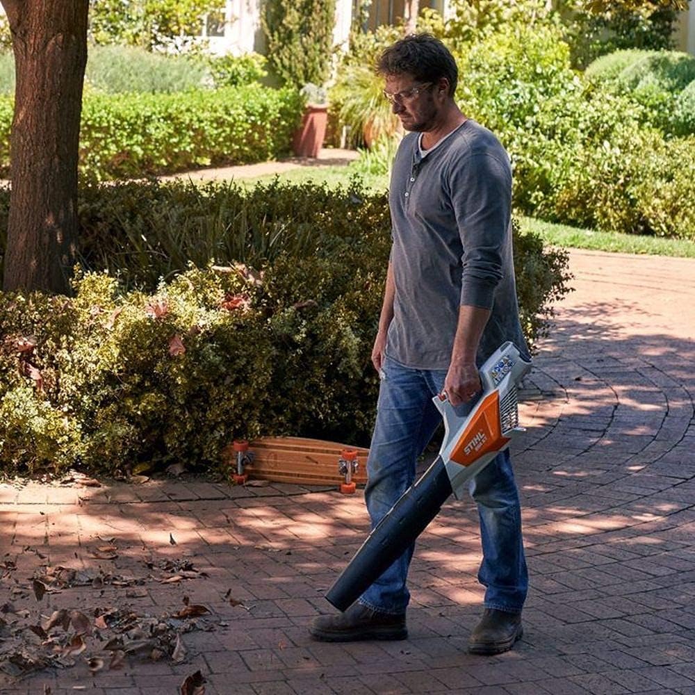 Uomo utilizza un soffiatore elettrico STIHL per pulire foglie secche su un vialetto di mattoni in giardino. Indossa occhiali, maglietta grigia e jeans. Sullo sfondo, cespugli verdi e una casa. Attrezzi da giardino visibili.