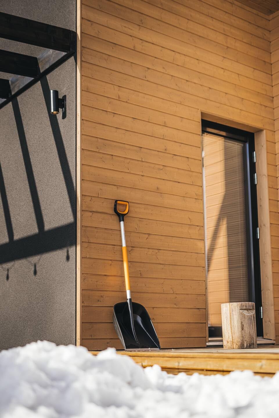 Pala da neve con manico arancione e nero appoggiata a una parete di legno chiaro accanto a una porta. Neve fresca in primo piano. Ideale per rimuovere la neve invernale. Strumento essenziale per l'inverno.