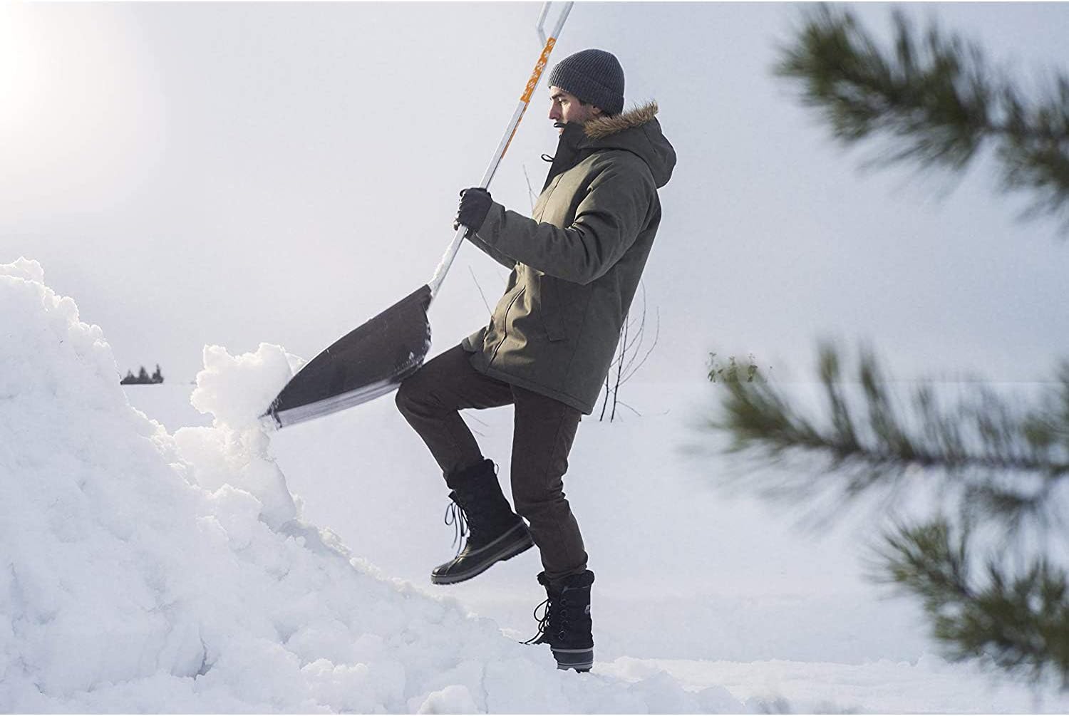 Uomo con giacca invernale, berretto e guanti spalando neve con una pala in un paesaggio innevato. Abbigliamento invernale, attrezzatura per spalare neve, attività all'aperto in inverno. Giacca con cappuccio, stivali invernali, comfort e calore.