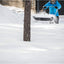 Persona con giacca blu che spala la neve in un cortile innevato, utilizzando una pala da neve a doppia impugnatura, con una casa in legno sullo sfondo. Strumenti per la rimozione della neve, inverno, attrezzatura da esterno.