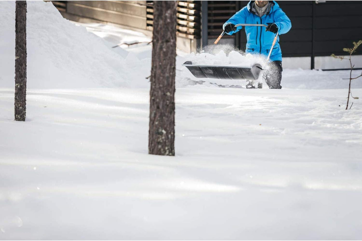 Persona con giacca blu che spala la neve in un cortile innevato, utilizzando una pala da neve a doppia impugnatura, con una casa in legno sullo sfondo. Strumenti per la rimozione della neve, inverno, attrezzatura da esterno.