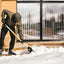 Persona che spalando neve fuori da una casa moderna in legno, indossando abbigliamento invernale: giacca verde, pantaloni neri, guanti e stivali da neve. Pala da neve con manico arancione. Sfondo con grandi finestre a vetri.