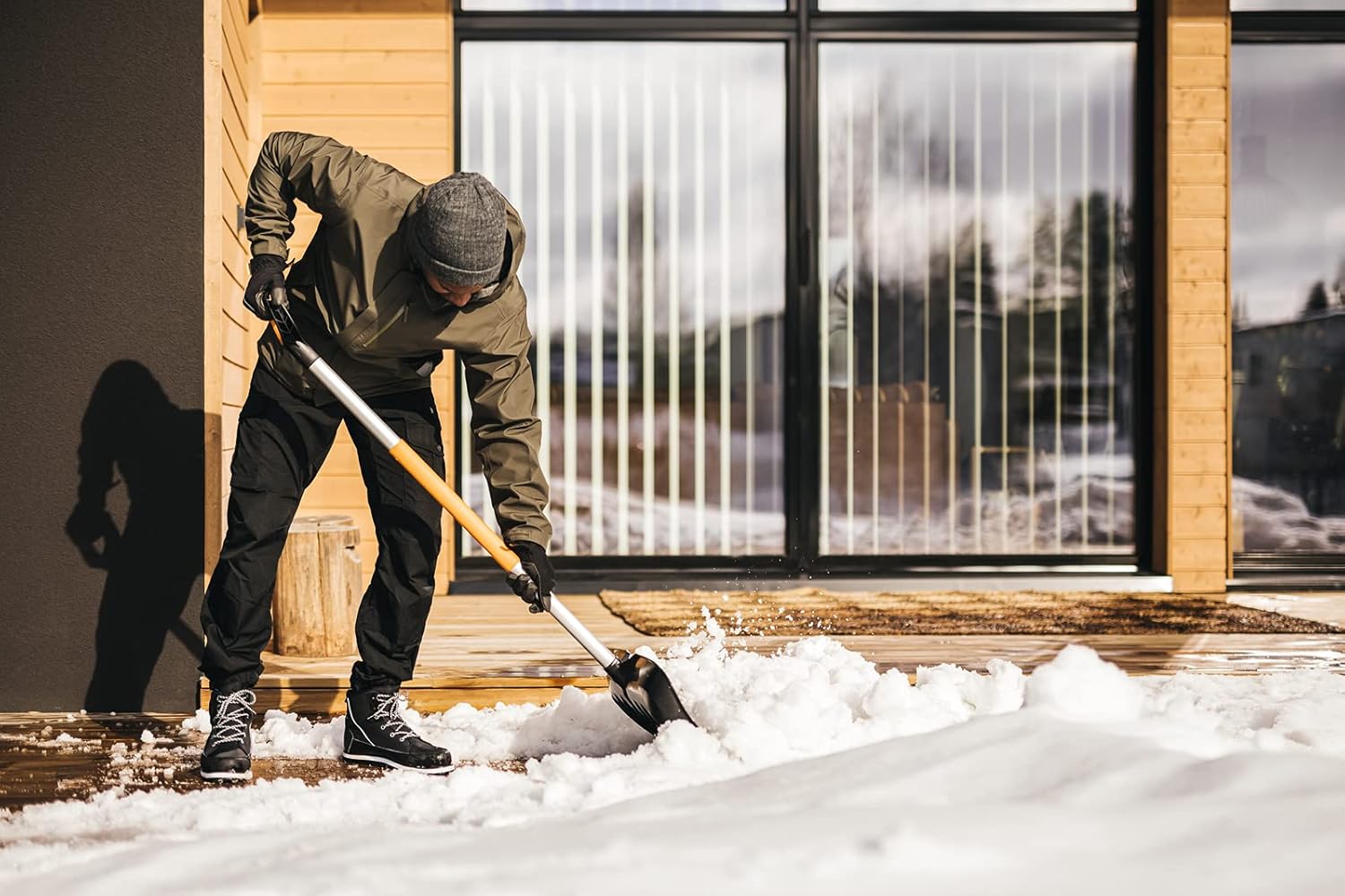 Persona che spalando neve fuori da una casa moderna in legno, indossando abbigliamento invernale: giacca verde, pantaloni neri, guanti e stivali da neve. Pala da neve con manico arancione. Sfondo con grandi finestre a vetri.