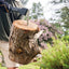 Persona che usa un'accetta Fiskars per tagliare un tronco di legno in un giardino verde. Indossa guanti protettivi e pantaloncini chiari. Attrezzo ideale per lavori di giardinaggio e taglio legna. Sfondo con alberi e fiori.