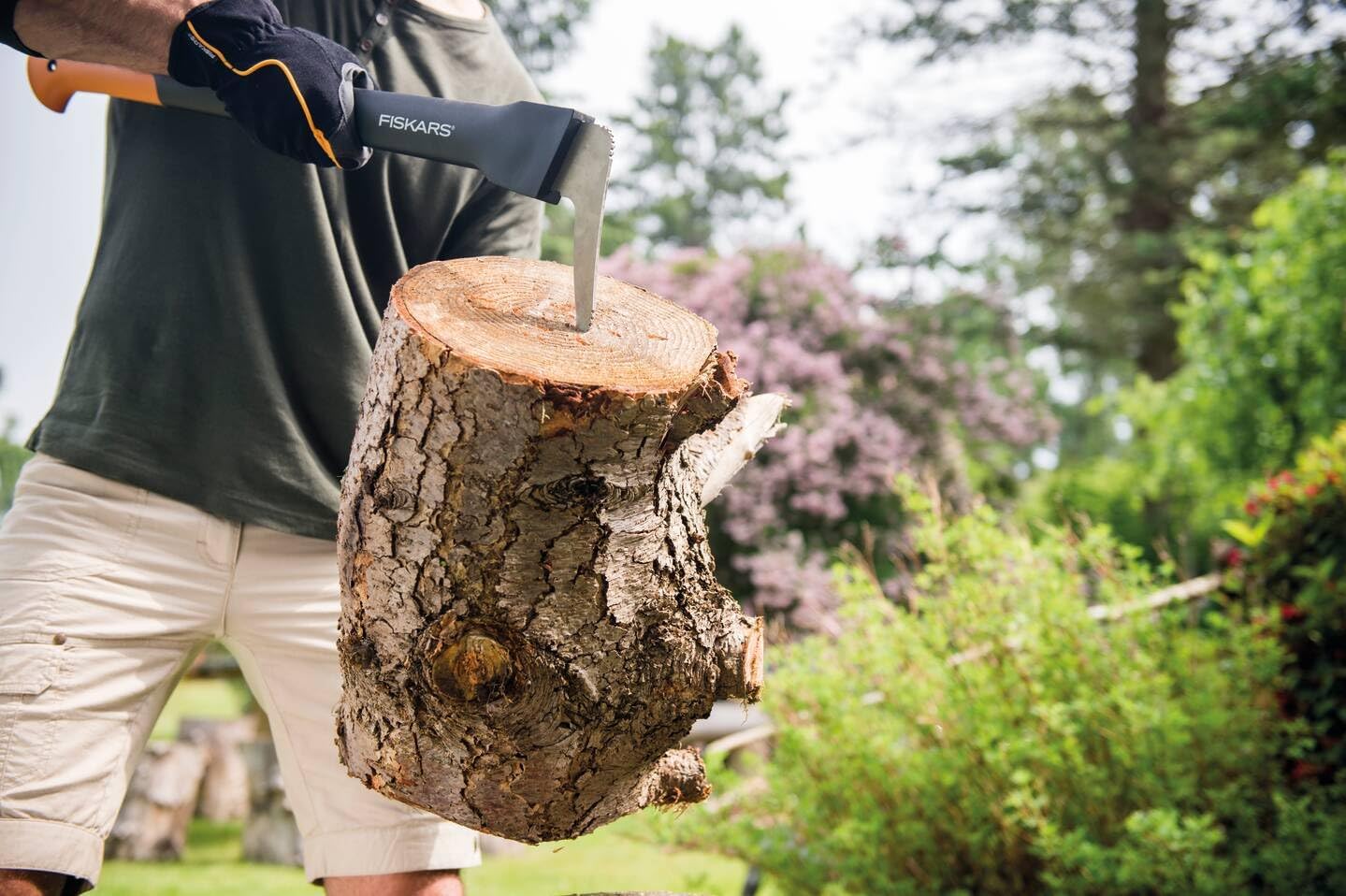 Persona che usa un'accetta Fiskars per tagliare un tronco di legno in un giardino verde. Indossa guanti protettivi e pantaloncini chiari. Attrezzo ideale per lavori di giardinaggio e taglio legna. Sfondo con alberi e fiori.