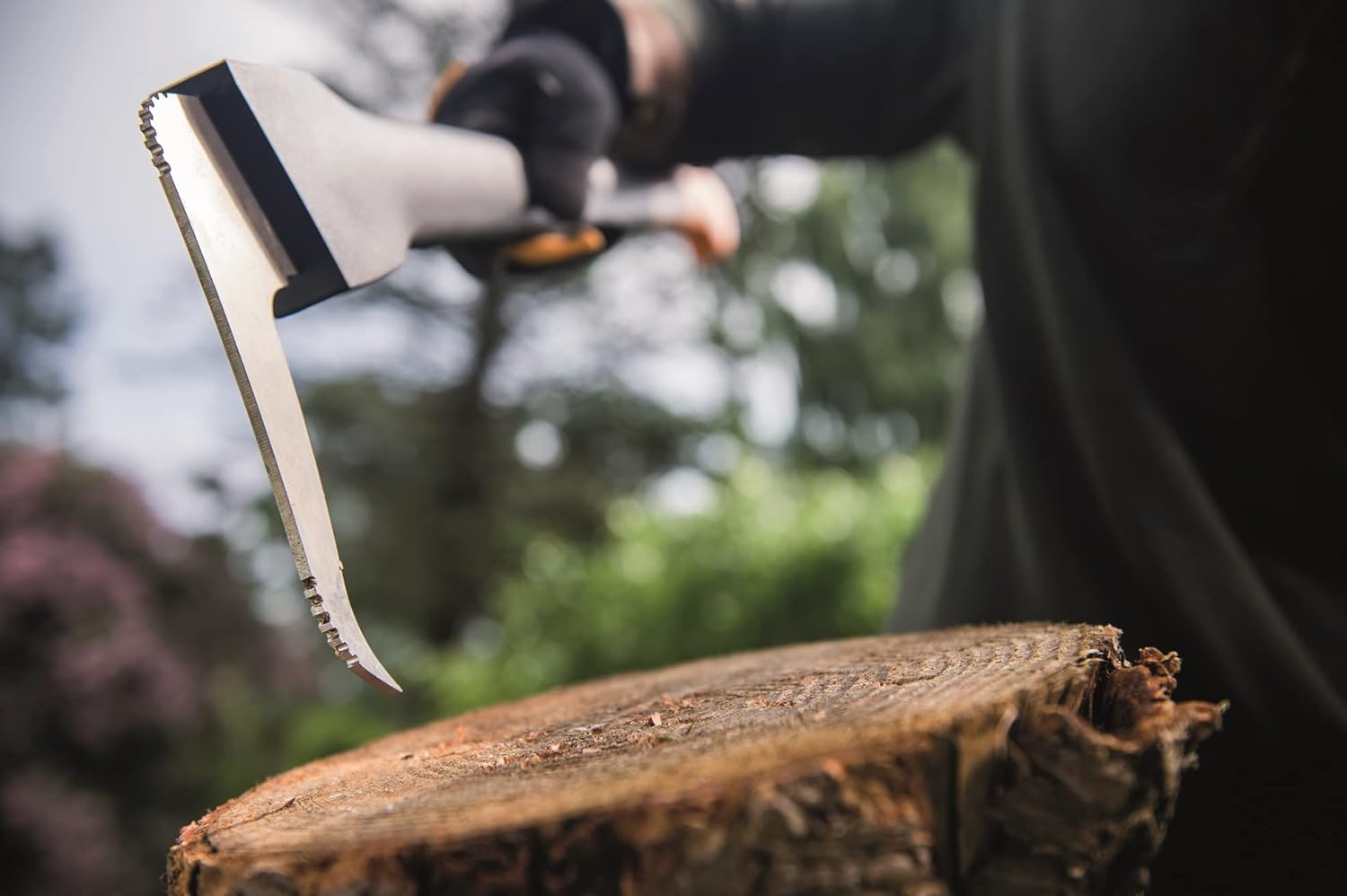 Primo piano di una scure con una lama affilata e dentellata che colpisce un ceppo di legno, utilizzata per tagliare legna in un ambiente esterno. Mano guantata che impugna la scure, sfondo sfocato con alberi e vegetazione.