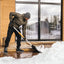 Uomo spalando neve su una veranda di legno davanti a una casa moderna con grandi finestre. Indossa abbigliamento invernale: giacca verde, pantaloni neri, guanti e berretto grigio. Scarpe invernali nere con suola robusta.