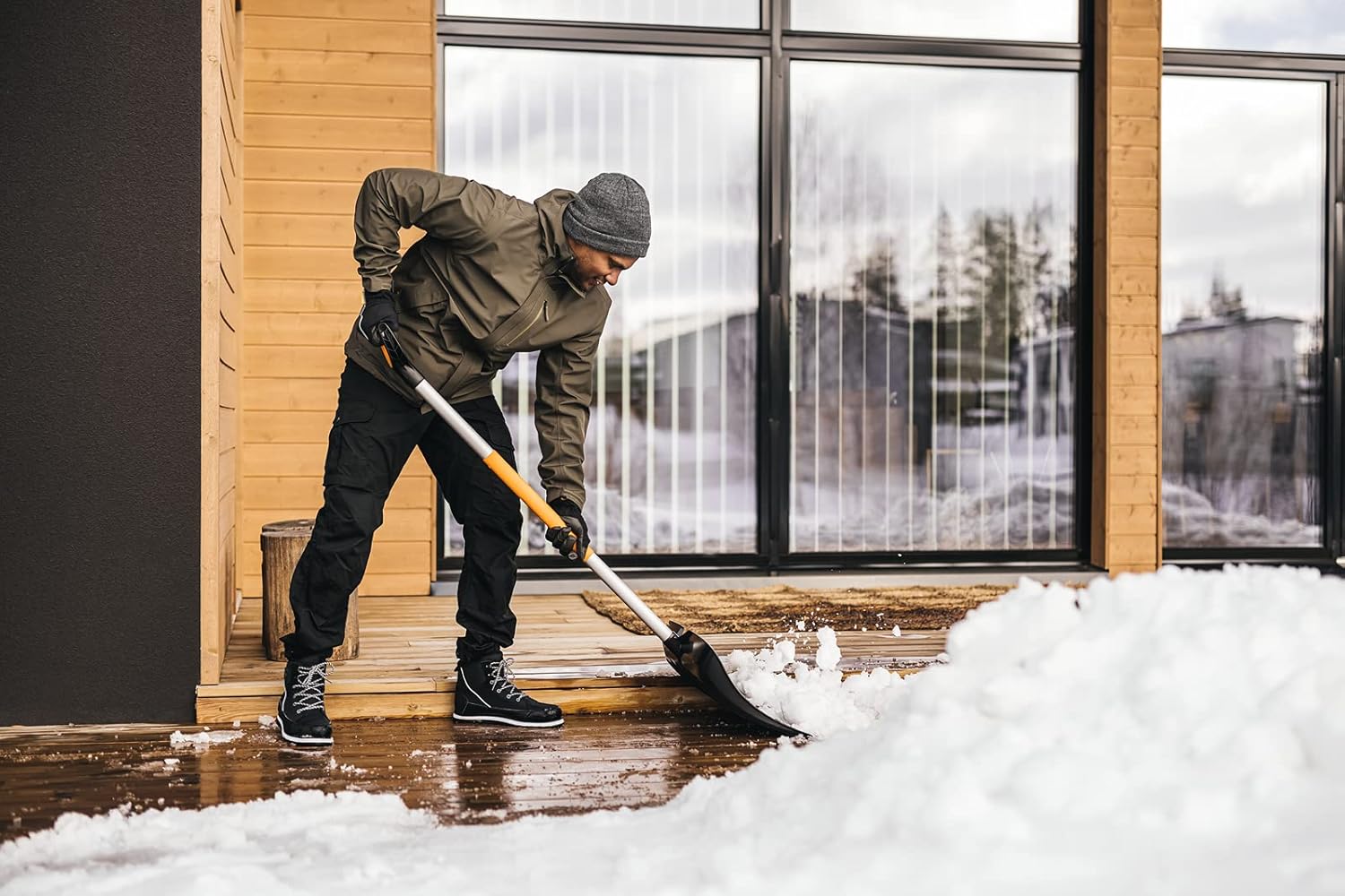 Uomo spalando neve su una veranda di legno davanti a una casa moderna con grandi finestre. Indossa abbigliamento invernale: giacca verde, pantaloni neri, guanti e berretto grigio. Scarpe invernali nere con suola robusta.