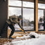 Uomo spalando neve su un patio in legno davanti a una casa moderna, indossando giacca invernale, pantaloni neri, guanti e berretto grigio. Scena invernale con attrezzo da neve, sfondo con finestre e paesaggio innevato.